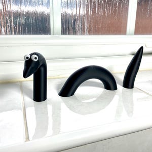 May include: A black, 3-piece, plastic, cartoon snake with googly eyes. The snake is made up of a head, a curved body, and a tail.
