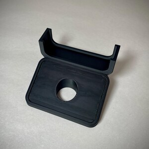 May include: A black, rectangular case with a circular opening and a hinged lid. The lid is open, revealing the interior of the case. The case is made of a textured material.