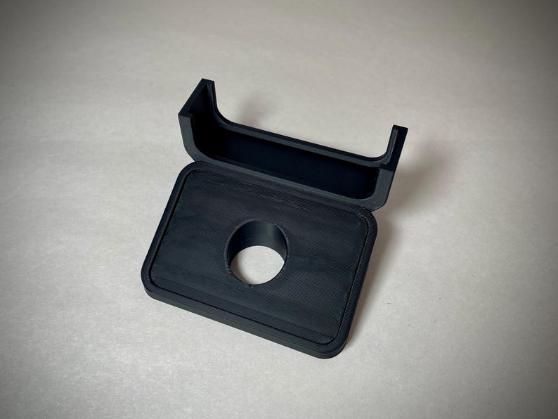 May include: A black, rectangular case with a circular opening and a hinged lid. The lid is open, revealing the interior of the case. The case is made of a textured material.