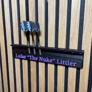 Puede incluir: Un soporte negro para dardos con tres dardos, montado en una pared de madera. Los dardos tienen ca&ntilde;as negras y plumas azules y negras. El soporte tiene el texto "Luke 'The Nuke' Littler" en morado.