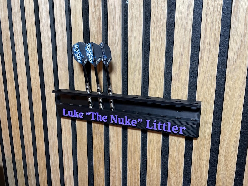 Puede incluir: Un soporte negro para dardos con tres dardos, montado en una pared de madera. Los dardos tienen ca&ntilde;as negras y plumas azules y negras. El soporte tiene el texto "Luke 'The Nuke' Littler" en morado.