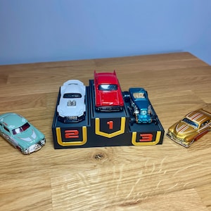 May include: A black three-tiered display stand with gold-outlined numbers 1, 2, and 3. A red car sits on the top tier, a white car on the second, and a teal car on the third. A light green car and a gold car are placed on either side of the stand.