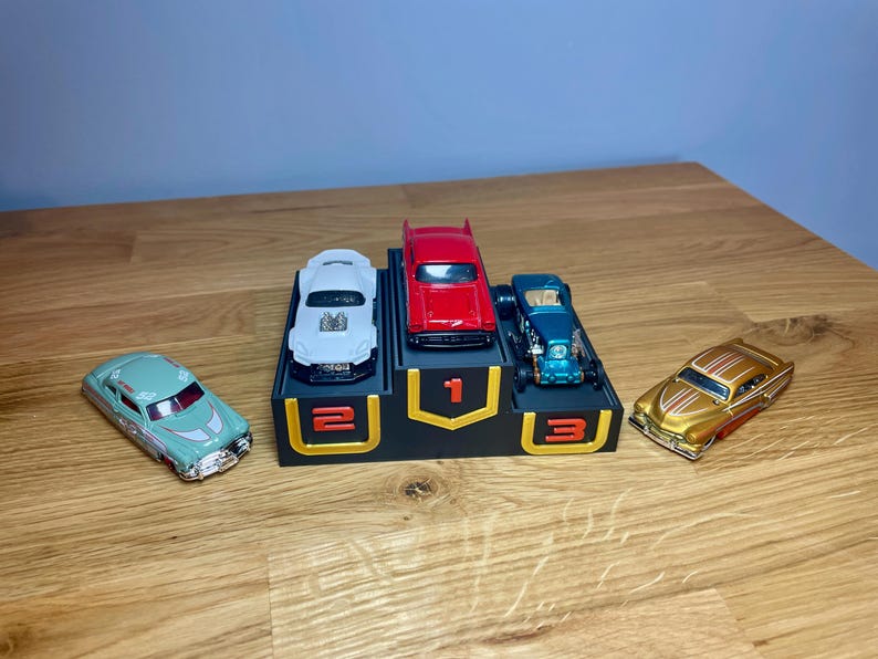 May include: A black three-tiered display stand with gold-outlined numbers 1, 2, and 3. A red car sits on the top tier, a white car on the second, and a teal car on the third. A light green car and a gold car are placed on either side of the stand.