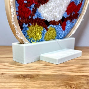 May include: A white wooden stand with a circular base and a rectangular platform, designed to display a colorful yarn-wrapped embroidery hoop.
