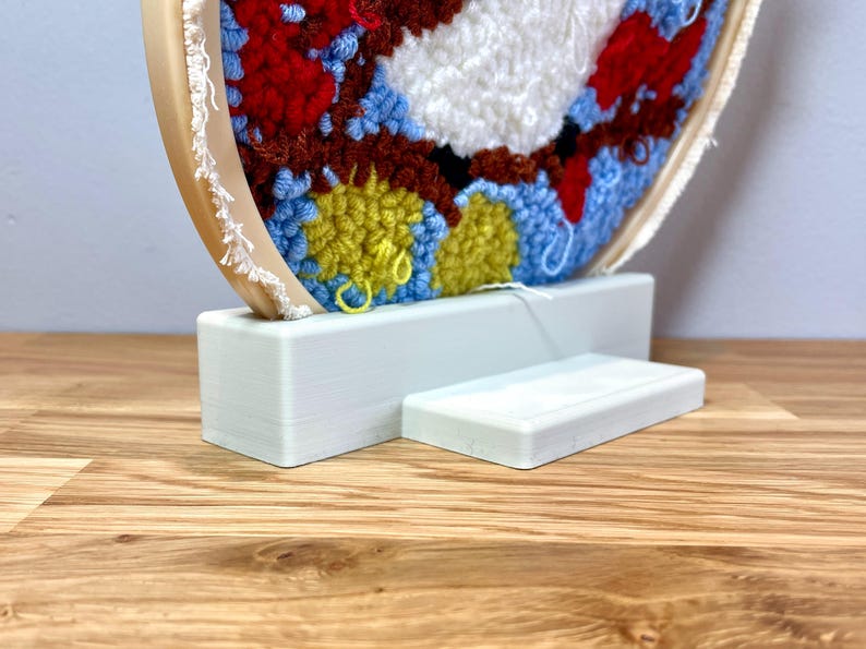 May include: A white wooden stand with a circular base and a rectangular platform, designed to display a colorful yarn-wrapped embroidery hoop.