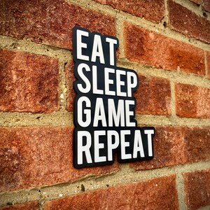 May include: A black and white sign displaying the text "EAT SLEEP GAME REPEAT" in a vertical arrangement. The sign is affixed to a red brick wall. The words are in a bold, sans-serif font.