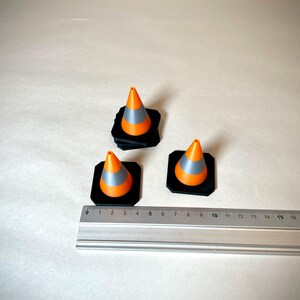 May include: Three miniature orange and grey traffic cones on black square bases. A metal ruler is placed near the cones, showing measurements in centimetres. The cones are arranged on a light-coloured surface.