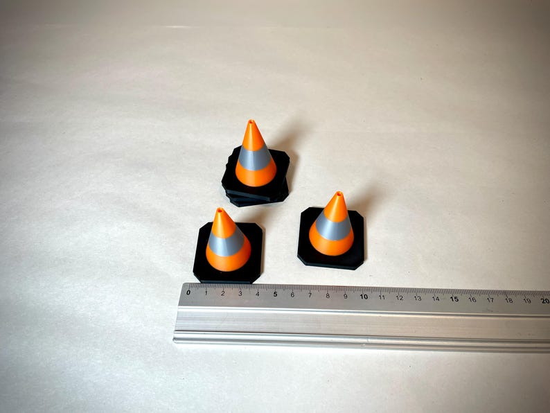 May include: Three miniature orange and grey traffic cones on black square bases. A metal ruler is placed near the cones, showing measurements in centimetres. The cones are arranged on a light-coloured surface.
