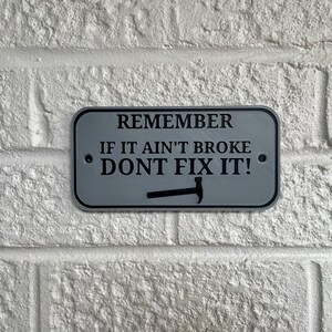 May include: A rectangular sign with rounded corners, featuring the text "REMEMBER IF IT AIN'T BROKE DON'T FIX IT!" in black letters. A hammer graphic is below the text. The sign is gray with a black border and is mounted on a white brick wall.