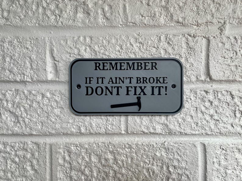 May include: A rectangular sign with rounded corners, featuring the text "REMEMBER IF IT AIN'T BROKE DON'T FIX IT!" in black letters. A hammer graphic is below the text. The sign is gray with a black border and is mounted on a white brick wall.