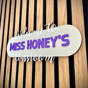 K&ouml;nnte beinhalten: Ein Klassenzimmerschild mit dem Text "Welcome to Miss Honey's classroom" in wei&szlig;er und violetter Schrift. Das Schild ist an einer Holzwand mit vertikalen Brettern befestigt.