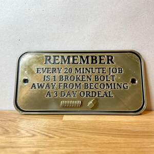 May include: A gold-colored metal sign with black text that reads "REMEMBER EVERY 20 MINUTE JOB IS 1 BROKEN BOLT AWAY FROM BECOMING A 3 DAY ORDEAL".