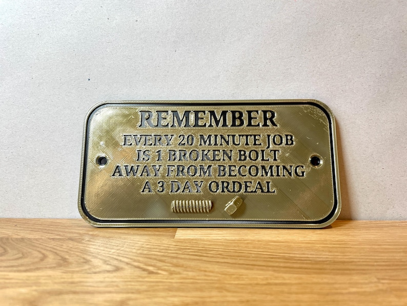 May include: A gold-colored metal sign with black text that reads "REMEMBER EVERY 20 MINUTE JOB IS 1 BROKEN BOLT AWAY FROM BECOMING A 3 DAY ORDEAL".
