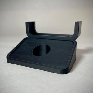 May include: A black, angled stand with a rectangular base and a curved top. The stand features a circular opening in the center of the base. The stand is made of a matte black material and is designed to hold an item.