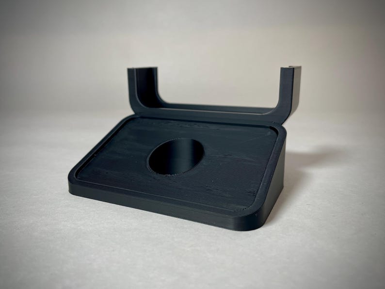 May include: A black, angled stand with a rectangular base and a curved top. The stand features a circular opening in the center of the base. The stand is made of a matte black material and is designed to hold an item.