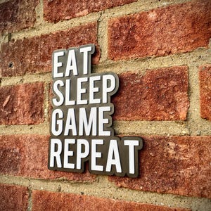 May include: A black and white sign with the words "EAT SLEEP GAME REPEAT" is mounted on a red brick wall. The sign has a white font with a black border. The brick wall has a rough texture.