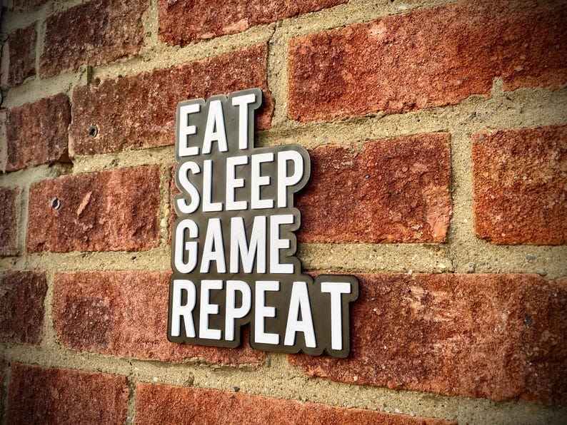 May include: A black and white sign with the words "EAT SLEEP GAME REPEAT" is mounted on a red brick wall. The sign has a white font with a black border. The brick wall has a rough texture.