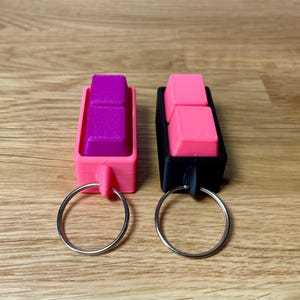 May include: Two keychains with pink and purple keycaps attached to silver key rings. The keychains are made of black and pink plastic.