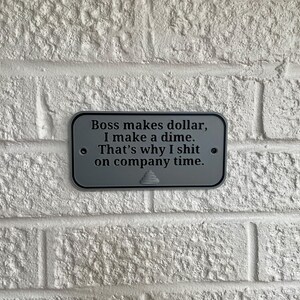May include: A rectangular, grey metal sign with the text "Boss makes dollar, I make a dime. That's why I shit on company time." The sign is mounted on a white brick wall.