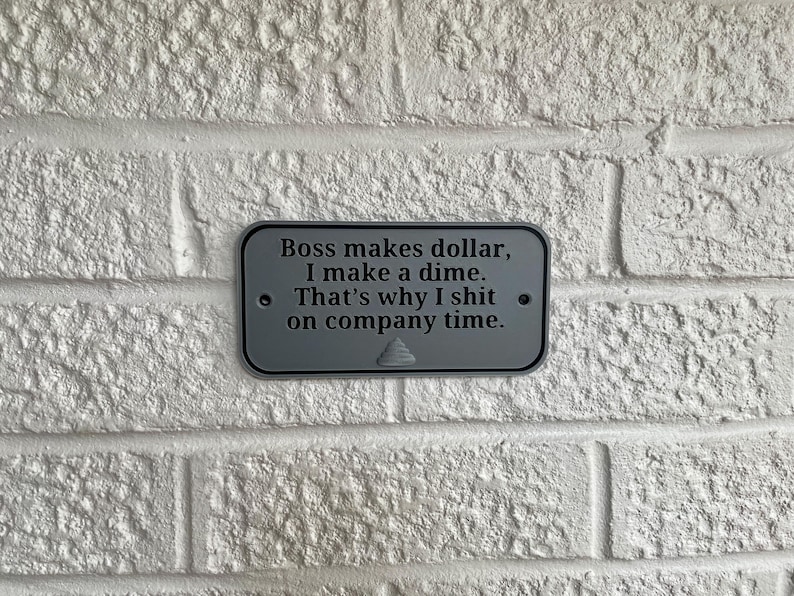 May include: A rectangular, grey metal sign with the text "Boss makes dollar, I make a dime. That's why I shit on company time." The sign is mounted on a white brick wall.