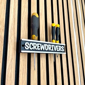 May include: A black and white screwdriver holder with the word "SCREWDRIVERS" in white lettering. The holder has three screwdrivers with yellow and black handles. The holder is mounted on a wooden wall with vertical stripes.