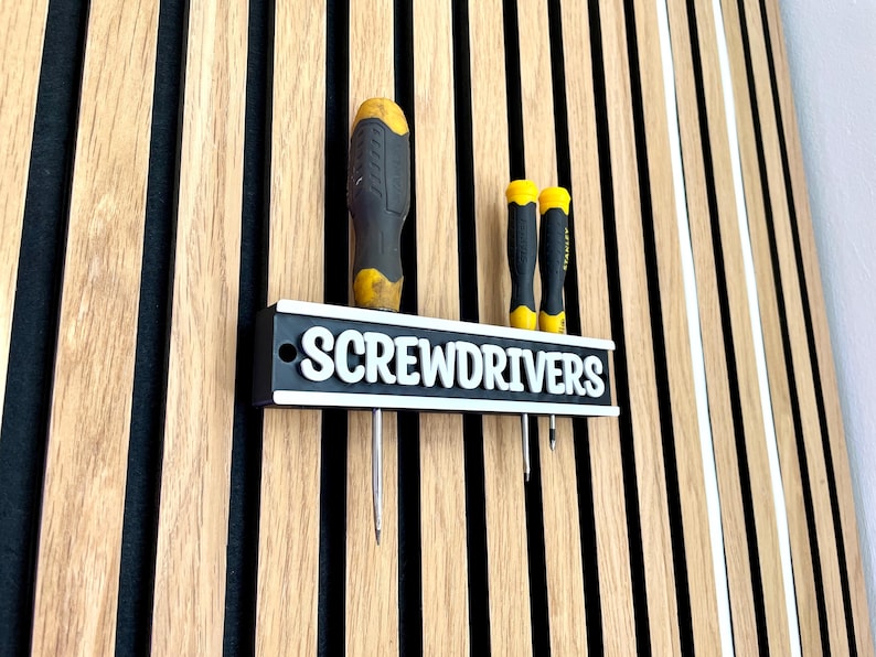 May include: A black and white screwdriver holder with the word "SCREWDRIVERS" in white lettering. The holder has three screwdrivers with yellow and black handles. The holder is mounted on a wooden wall with vertical stripes.