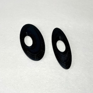 May include: Two black, oval-shaped earrings with a central hole. The earrings have a textured surface and are displayed on a white background. The design is simple and modern, suitable for everyday wear.