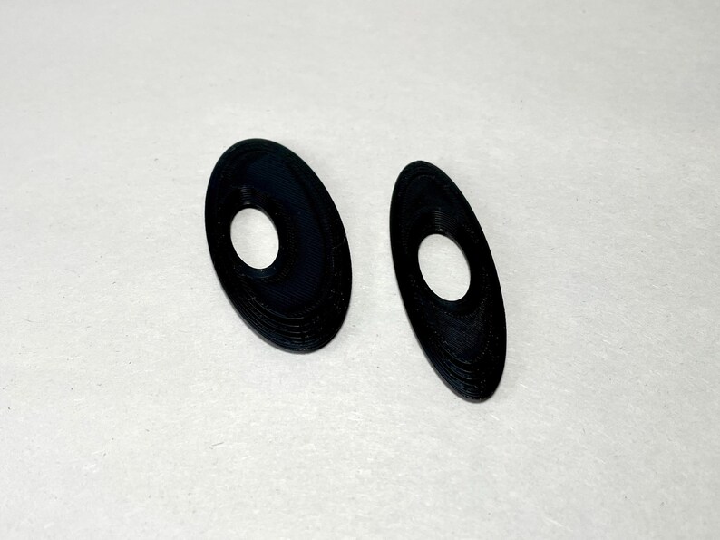 May include: Two black, oval-shaped earrings with a central hole. The earrings have a textured surface and are displayed on a white background. The design is simple and modern, suitable for everyday wear.