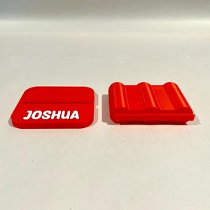 Op de afbeelding: Twee rode, 3D-geprinte items. E&eacute;n is een rechthoekige vorm met de naam "JOSHUA" in witte letters. De andere is een rood, geribbeld, rechthoekig object. Beide items liggen op een wit oppervlak.