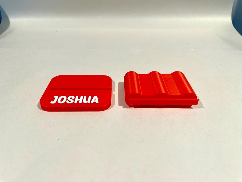Op de afbeelding: Twee rode, 3D-geprinte items. E&eacute;n is een rechthoekige vorm met de naam "JOSHUA" in witte letters. De andere is een rood, geribbeld, rechthoekig object. Beide items liggen op een wit oppervlak.