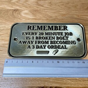 May include: A silver metal sign with black text that reads "REMEMBER EVERY 20 MINUTE JOB IS 1 BROKEN BOLT AWAY FROM BECOMING A 3 DAY ORDEAL".