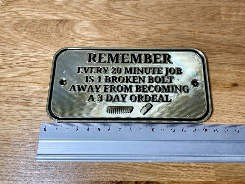 May include: A silver metal sign with black text that reads "REMEMBER EVERY 20 MINUTE JOB IS 1 BROKEN BOLT AWAY FROM BECOMING A 3 DAY ORDEAL".