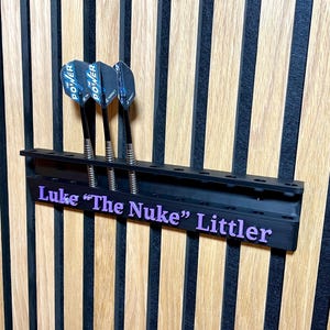 Puede incluir: Soporte negro para dardos con tres dardos. Los dardos tienen ca&ntilde;as negras y plumas Power azules y negras. El soporte tiene el nombre "Luke "The Nuke" Littler" en texto morado.