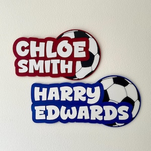 May include: Two soccer-themed name tags. One is burgundy with the name "CHLOE SMITH" in white, and the other is blue with "HARRY EDWARDS" in white. Each tag has a soccer ball design.