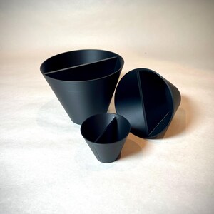May include: Three black, cone-shaped containers of varying sizes. Each container is divided into two sections by a vertical divider. The containers are arranged on a light-colored surface, with the largest one in the background.