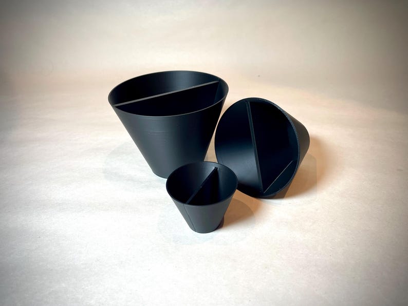May include: Three black, cone-shaped containers of varying sizes. Each container is divided into two sections by a vertical divider. The containers are arranged on a light-colored surface, with the largest one in the background.