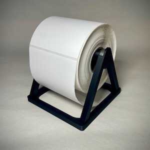 May include: A roll of white labels sits on a black, triangular label holder. The label roll is large and cylindrical, with a visible seam. The holder is made of a dark material and is designed to keep the labels organized.