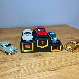 May include: A black three-tiered display stand with yellow accents and the numbers 1, 2, and 3 in orange. A red toy car sits on the top tier, a white toy car on the second, and a teal toy car on the third. A light green and a gold toy car sit nearby.