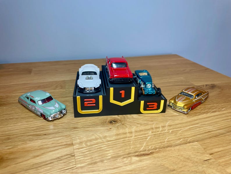 May include: A black three-tiered display stand with yellow accents and the numbers 1, 2, and 3 in orange. A red toy car sits on the top tier, a white toy car on the second, and a teal toy car on the third. A light green and a gold toy car sit nearby.