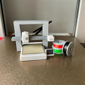 Pu&ograve; includere: Una stampante 3D con telaio grigio e componenti bianchi, tra cui un piano di stampa e bobine di filamento rosso, verde e nero. La stampante ha il testo "A1" su uno dei suoi componenti.