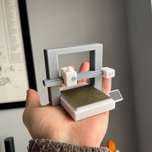 Pu&ograve; includere: Un piccolo modello di stampante 3D grigio e bianco con un piano di stampa color oro. Il modello &egrave; tenuto in mano, mostrando le sue dimensioni compatte e il design. La stampante ha un telaio rettangolare e una piccola testina bianca.
