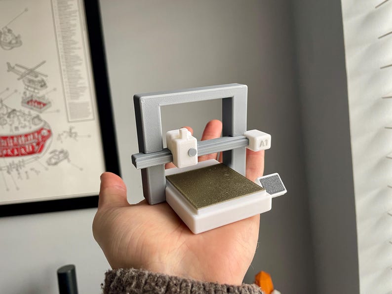 Pu&ograve; includere: Un piccolo modello di stampante 3D grigio e bianco con un piano di stampa color oro. Il modello &egrave; tenuto in mano, mostrando le sue dimensioni compatte e il design. La stampante ha un telaio rettangolare e una piccola testina bianca.