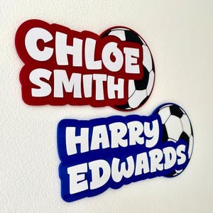 May include: Two personalized name tags with soccer ball accents. One is red with "CHLOE SMITH" in white, the other is blue with "HARRY EDWARDS" in white. Both have a soccer ball design.