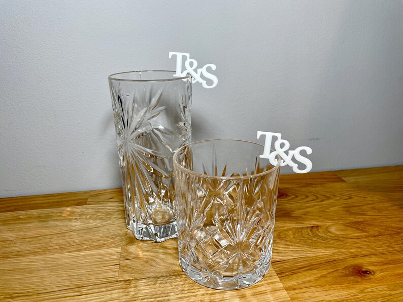 Tumbler and tall glass with a set of initial letters clipped onto the side. Drink topper is white and the photo shows a grey background.