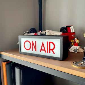 May include: A black rectangular "ON AIR" sign with red lettering, illuminated against a white background. The sign is on a wooden shelf with other items, including a Lego race car and a trophy.