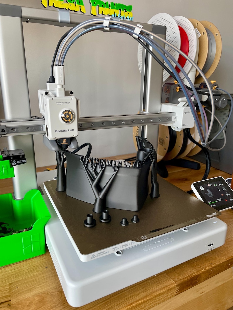 May include: A 3D printer with a white base and silver frame. The printer has a black object on the print bed and several spools of filament in the background. The brand name "Bambu Lab" is visible on the printer.