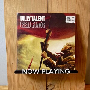 May include: A vinyl record by Billy Talent titled "Red Flag" with a red, white, and blue flag design and a person holding the flag. The record is on a wooden shelf with a black sign that says "NOW PLAYING".