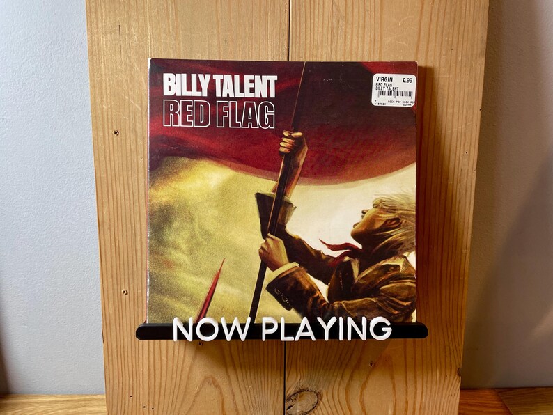 May include: A vinyl record by Billy Talent titled "Red Flag" with a red, white, and blue flag design and a person holding the flag. The record is on a wooden shelf with a black sign that says "NOW PLAYING".