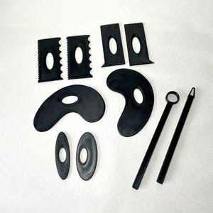 May include: A collection of black pottery tools, including various shaped ribs, loop tools, and a needle tool. The tools are arranged on a white surface, showcasing their different shapes and sizes. These tools are used for shaping and smoothing clay.