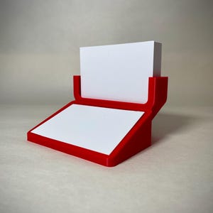 May include: A red and white business card holder. The holder has a red frame with a white surface for the cards and a white block to hold the cards in place. The design is modern and minimalist.